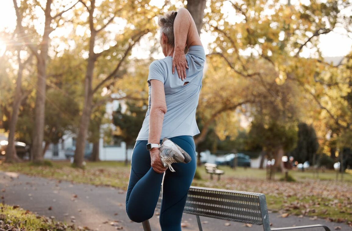 An active senior woman seen from behind stretching her arm and leg in a sunlit park.